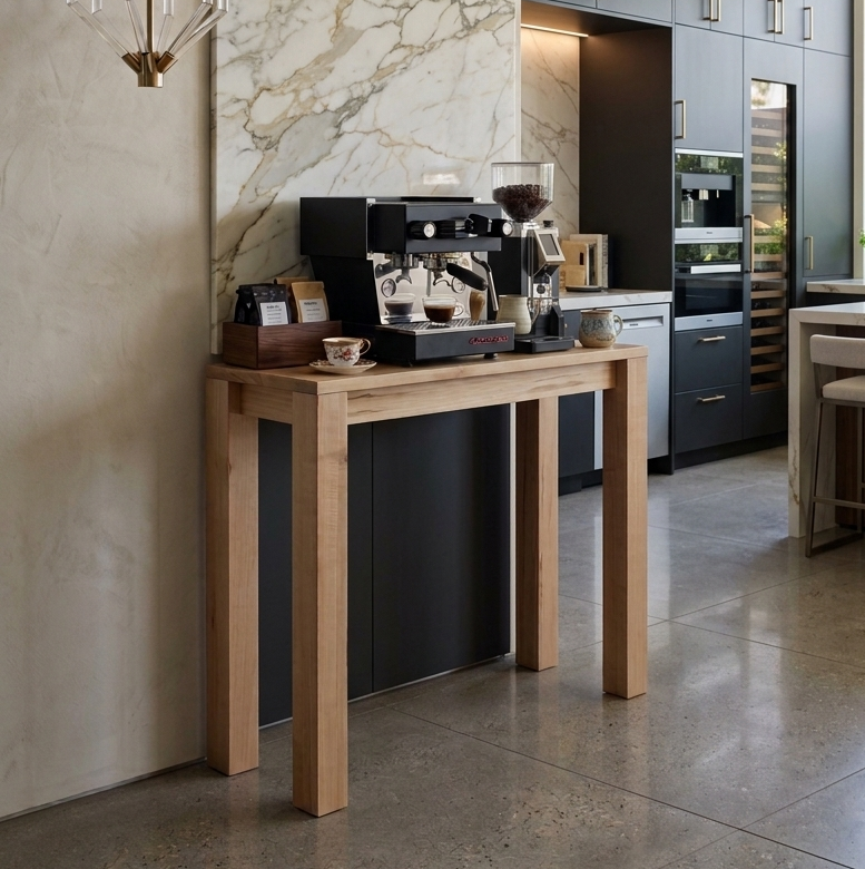 A modern kitchen interior with a light wood console table holding a black espresso machine, set against a large beige marble wall. Dark charcoal kitchen cabinets, built-in appliances, a hanging glass chandelier, and a kitchen island with bar stools are visible in the background.